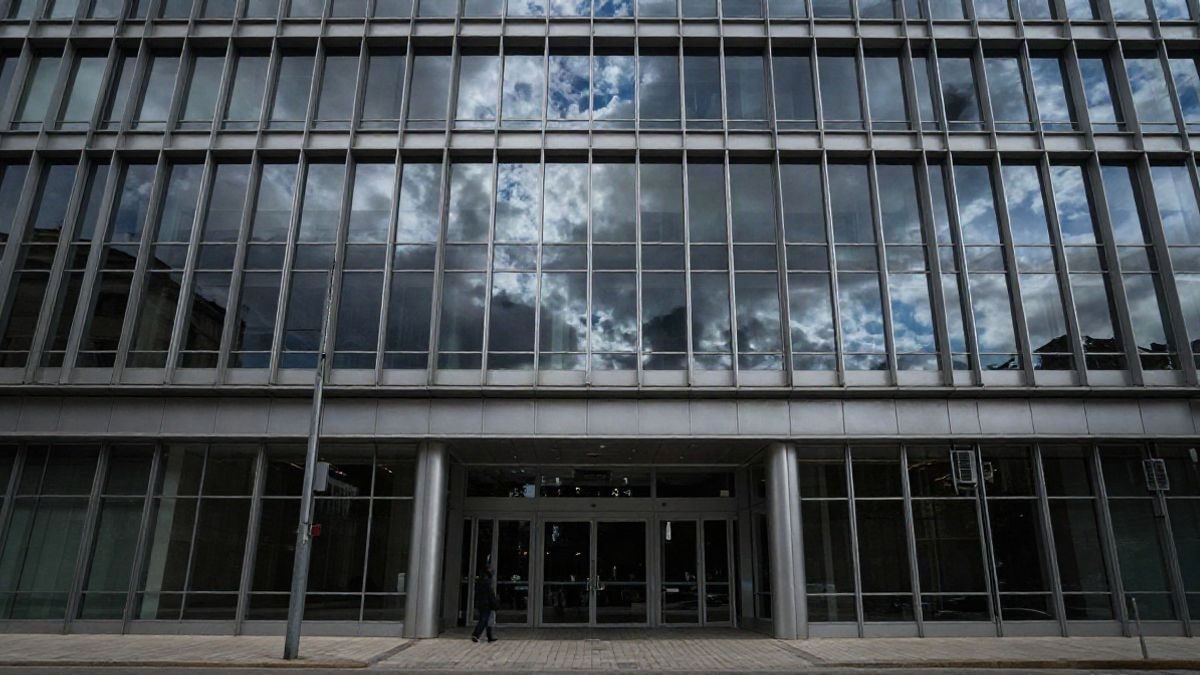 Imagen ilustrativa: fachada de un edificio de oficinas moderno con grandes ventanales reflejando el cielo nublado, con una figura humana lejana y desenfocada